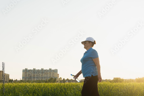 Active senior woman in sport clothes hiking on outdoor in the field with bottle of water. Summer season. Selective focus. Wellness, healthy lifestyle