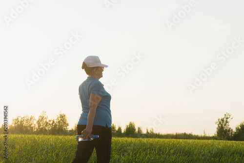Active senior woman in sport clothes hiking on outdoor in the field with bottle of water. Summer season. Selective focus. Wellness, healthy lifestyle