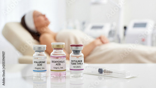 Three vials of Hyaluronic Acid, Deoxycholic Acid, and Botulinum Toxin Type A for anti-aging injections on a white table in a medical spa salon with a blurred female patient in the background.