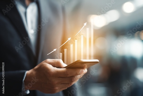 Business professional using a smartphone to analyze financial growth with upward arrows and bar graph indicating success in modern office environment