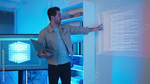 A professional male software engineer stands presenting and explaining lines of code projected on a wall display during a technical meeting, gesturing towards the data script in a dark room with drama