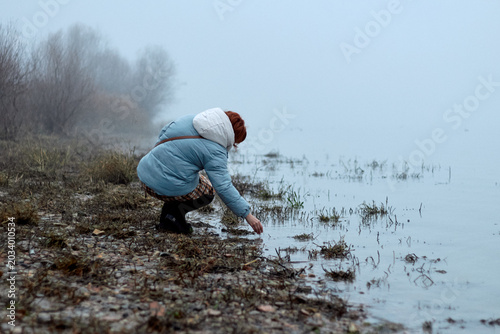 Woman crouching by misty lakeshore touching water in cold autumn morning