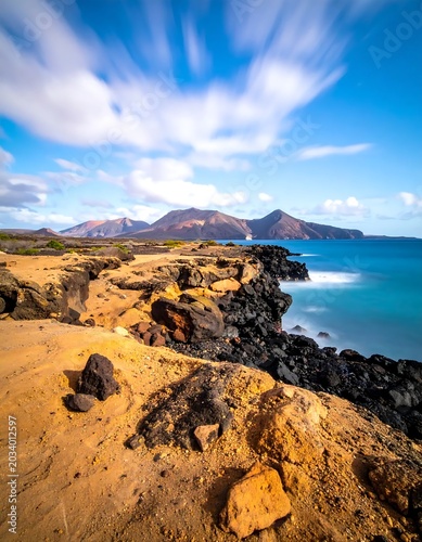 Dramatic Volcanic Coastline Overlooking Turquoise Ocean and Distant Mountains Under Cloudy Blue Sky