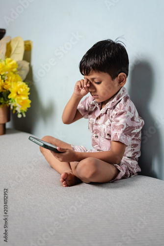 Young Indian boy sitting cross-legged, rubbing tired eyes while looking at a mobile phone screen