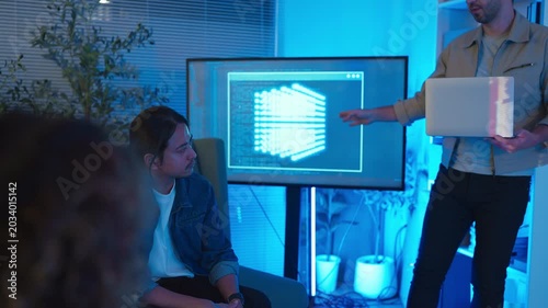 A professional East Asian female software engineer stands holding a laptop and gesturing toward lines of code projected on a wall display, presenting data scripts to her multiethnic colleagues during 