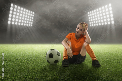An exhausted Indonesian southeast asian female football or soccer player in an orange uniform sits on the turf under bright stadium floodlights, looking dejected and disappointed after a game