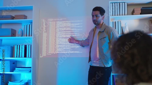 A professional male software engineer stands presenting and explaining lines of code projected on a wall display during a technical meeting, gesturing towards the data script in a dark room with drama