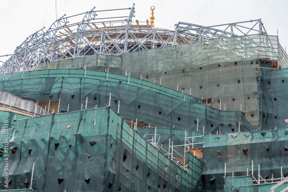 Fototapeta premium Construction of a large building in an urban area during cloudy weather with scaffolding and green netting present