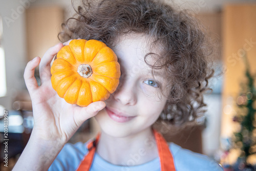 Playful smiling person holds two mini pumpkins over their eyes like glasses. Wearing an orange apron in a cozy kitchen, this fun, warm image is perfect for cheerful autumn and Halloween marketing.