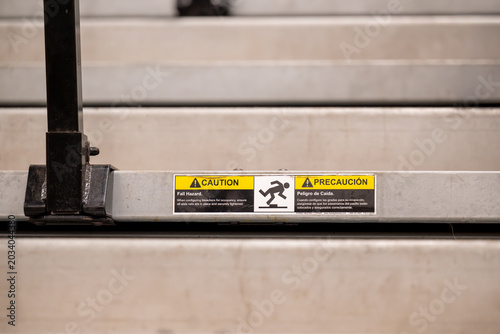 Canvastavla A bilingual English and Spanish caution label warning of fall hazard is affixed to the metal edge of bleacher steps, its yellow warning triangles and falling figure icon clearly legible
