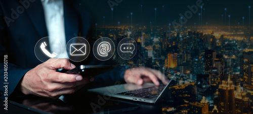 Businessman using smartphone and laptop with contact icons for customer support, email communication and digital networking in smart city technology and online business concept.