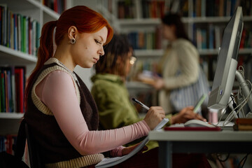 Woman studying at desktop computer, taking notes in library with classmates nearby. Education concept for academic success, exam prep, digital learning campaigns
