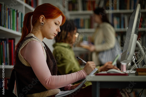 Woman studying at desktop computer, taking notes in library with classmates nearby. Education concept for academic success, exam prep, digital learning campaigns
