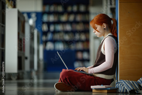 Female student studying on laptop while sitting on library floor, focusing on online learning and academic research
