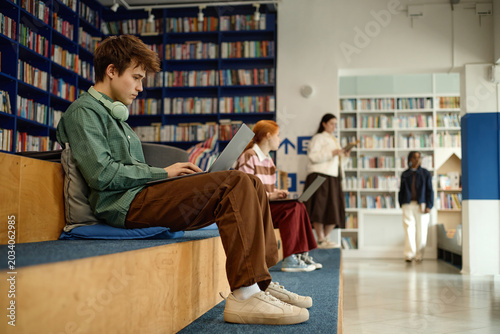 Male student studying on laptop in library, wearing headphones and preparing coursework while classmates learning nearby. Useful for education marketing, e learning, student life
