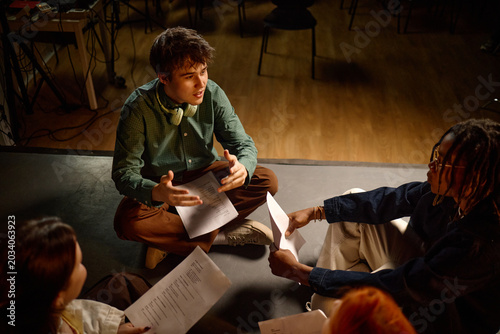 High angle shot of diverse students sitting on floor, discussing scripts during class project rehearsal
