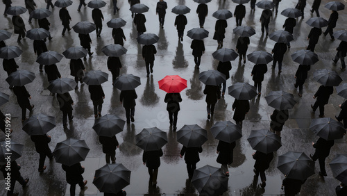 Crowd with black umbrellas on rainy city street, one red umbrella stands out. Concept of individuality, leadership, uniqueness, contrast, standing out from the crowd, urban lifestyle.