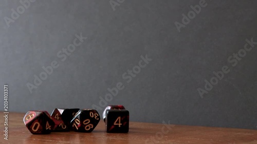 A pile of different dice like a D6, percentile dice, D4, D10, D12 and D8 sit on a wooden table. A player rolls a D20 into frame and after a few seconds picks it back up.