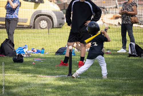 Young child swings bat at baseball field. Green grass covers entire playing surface. Adults stand behind netting, watching game. Equipment scattered nearby, including helmets.
