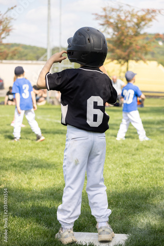 Young boy in black baseball jersey and helmet with white pants stands on base. Other players in blue jerseys stand nearby.