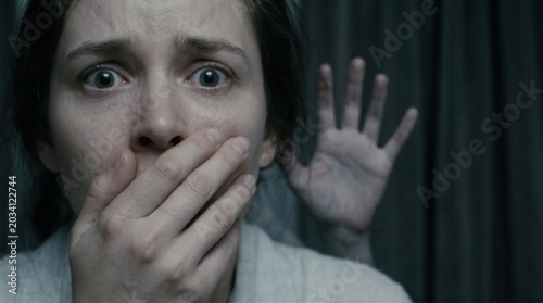 terrified young woman covers her mouth in extreme fear with a mysterious blurry ghost hand on dark background