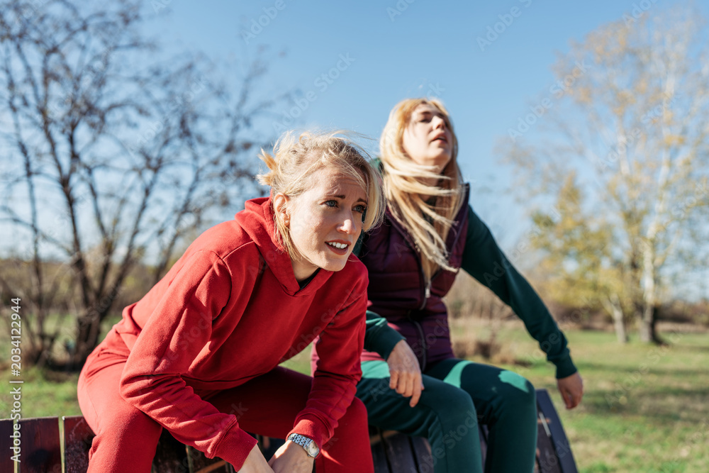 Naklejka premium Two tired female friends in sportswear resting on a wooden bench, catching their breath after an intense outdoor running workout in a sunny park.