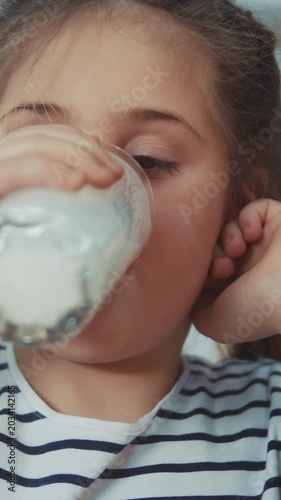 A girl drinks milk from a glass, focusing on the act of drinking milk. Her hand supports her face as she enjoys the creamy milk, surrounded by a light-setting.