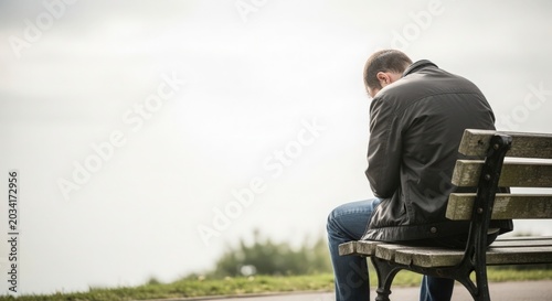 Man sitting alone on bench looking down in reflection during cloudy day. Emotional and contemplative moment captures a man lost in thought. Concept of solitude and inner struggle on a dreary day.