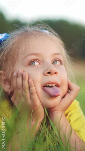 A girl is lying in the grass, gazing upwards with her head resting on her hands. The girl has bright eyes and a playful expression, adding to the serene outdoor scene.