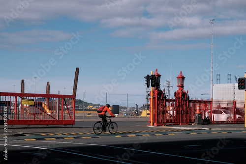 Auckland, New Zealand: a male urban cyclist rides past striking red industrial maritime gate barriers near the downtown ferry terminal on a sunny day.
