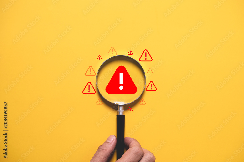Fototapeta premium Warning alert concept with magnifying glass focusing on danger symbol over yellow background. Risk management, security monitoring, problem detection, caution and safety awareness concept.