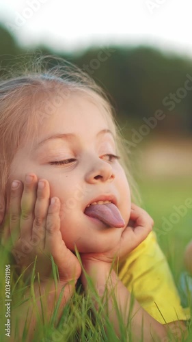 A girl is lying in the grass, resting her chin on her hands and sticking out her tongue. The girl appears to be enjoying a sunny day in a green outdoor setting.