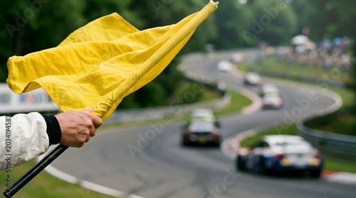 Hand holding a yellow flag on a race track with blurred racing cars in background.