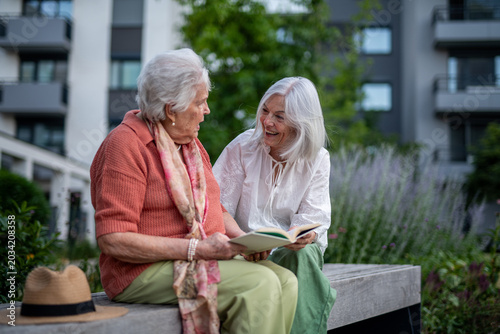Konstfotografi Elderly mother sitting on bench with adult daughter, reading together