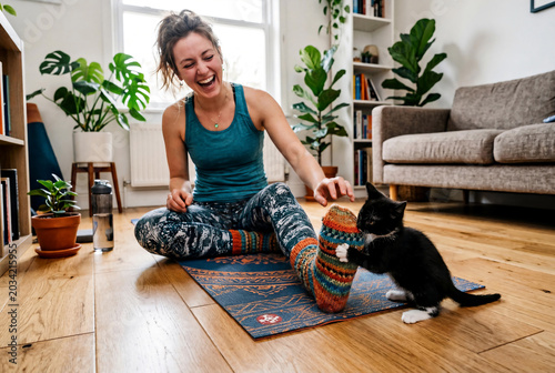 Laughing young woman interrupted by funny black kitten biting her colorful wool sock during yoga practice on mat at home for pet owner lifestyle or cozy wellness concept
