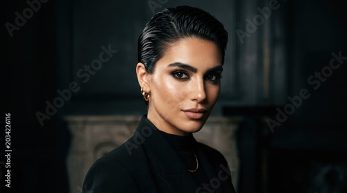 A woman with dark hair and makeup, wearing a black outfit, standing in a dimly lit room with a dark background.