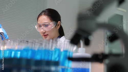 Asian female researcher wearing safety goggles and using a pipette to transfer blue liquid into test tubes during a scientific experiment.