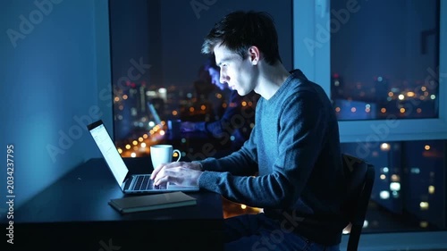 White man on laptop at night by window with city lights coffee mug and notebook beside him typing code under blue ambient glow reflective glass portrait conveying focused solitude and remote developer