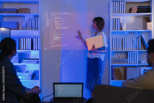 A professional East Asian female software engineer stands holding a laptop and gesturing toward lines of code projected on a wall display, presenting data scripts to her multiethnic colleagues during 