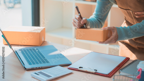 A young female entrepreneur checks order details on her smartphone and laptop, displaying a cardboard box. This is an online shopping business idea for small and medium-sized enterprises (SMEs).