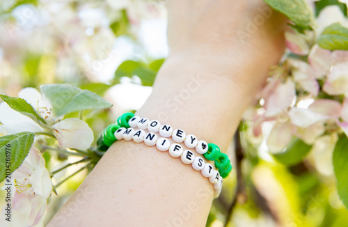 A person wearing a beaded bracelet made of letter beads spelling: money manifestation. Apple tree blossoms in the background, with beautiful bokeh lighting and a soft, dreamy backdrop. 