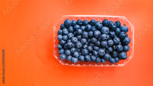 Fresh blueberries filling a clear plastic punnet, offering a vibrant contrast against the solid orange background, highlighting concepts of healthy eating and fresh produce