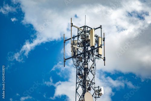 Telecommunications cell tower under blue sky with clouds. Low angle view of a modern telecommunication antenna tower showcasing wireless technology against a bright blue sky with puffy white clouds.