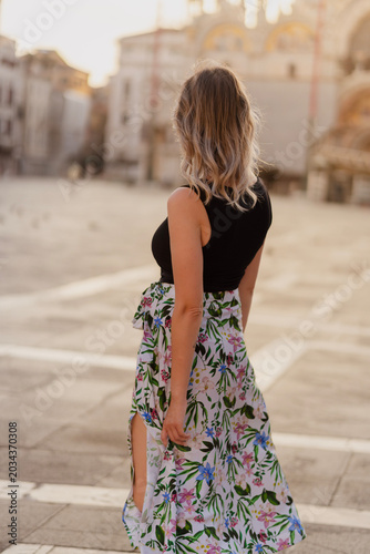 Elegant young woman from behind in a black tank top and floral skirt standing in the iconic St. Mark's Square in Venice, Italy at warm golden sunset