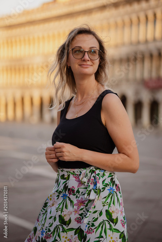 Stylish young woman with glasses in a black tank top and floral skirt posing at the iconic St. Mark's Square in Venice, Italy at warm golden sunset