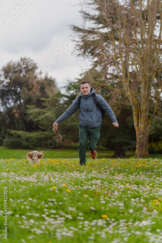 Athletic young man running and playing with his fluffy Cavalier King Charles Spaniel dog on a green wildflower meadow in a park on a cloudy spring day