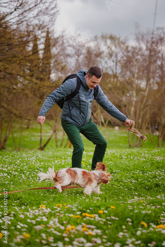 Young man playing and having fun with his fluffy dog on a green meadow with wildflowers in a park on a cloudy spring day