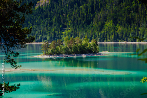 Panoramic view of a crystal-clear turquoise alpine lake with a forested island framed by pine tree branches and steep mountain slopes in summer