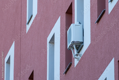 Modern pink building with white geometric window frames. Outdoor air conditioning unit mounted on wall. Minimalist urban architecture, clean lines, geometric pattern. Suitable for real estate