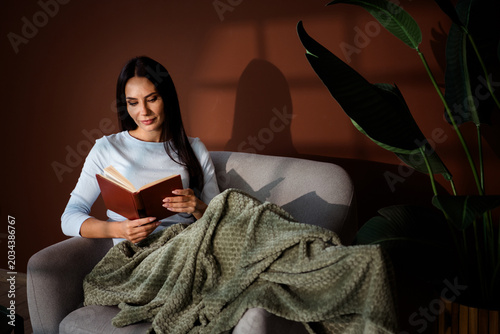 Woman reading a book on a sofa wrapped in a blanket in warm window light, relaxed at home with houseplant and cozy interior